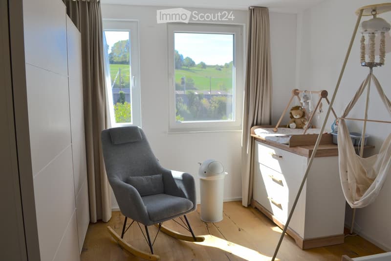 Brightly lit bedroom with large windows, a rocking chair, a baby crib with a swing, and a wardrobe.