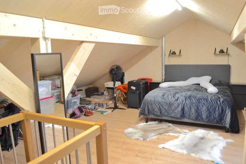 Attic bedroom with wooden floors, slanted ceilings, and exposed beams. Features a large bed with grey bedding, nightstand, fan, suitcase, and a mirror on the wall.