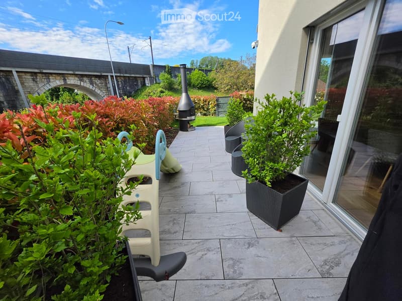 A well-maintained outdoor patio with a tiled floor, potted plants, a small table and chairs, and a fireplace in the background.