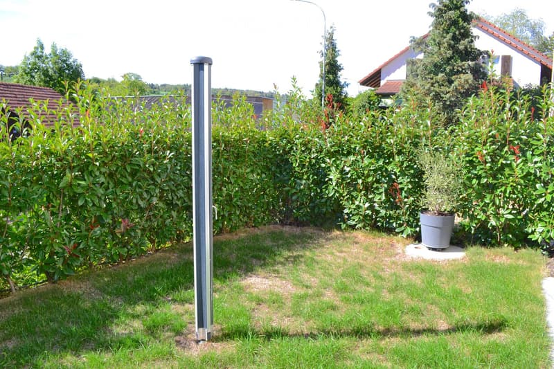 A well-maintained garden with a neatly trimmed hedge, a potted plant, and a modern streetlight pole. The lawn is green with patches of grass and dirt, and residential houses are visible in the background.