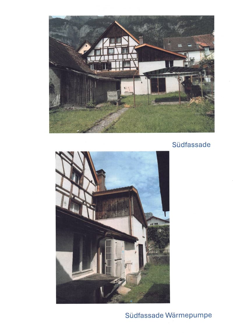 Traditional half-timbered houses with pitched roofs, surrounded by grassy areas and mountains in the background.