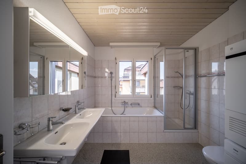Modern bathroom with a double sink, bathtub, shower, and large windows providing natural light. Features include a mirrored cabinet, tiled walls, and a sleek design.