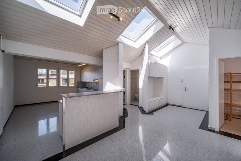 Bright, open loft-style room with white walls, wooden ceiling beams, skylights, and a modern kitchen area with cabinets and a countertop.