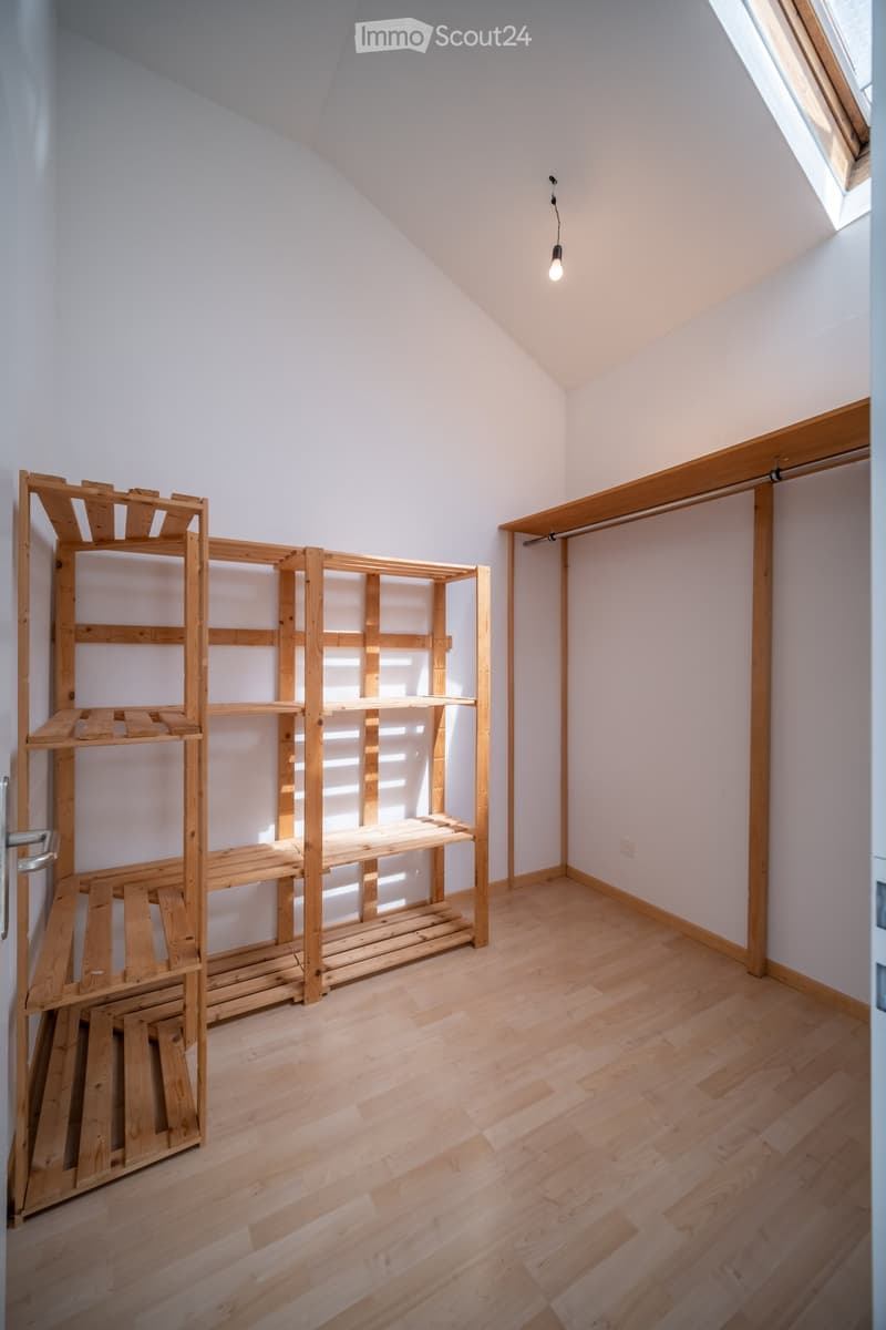 Empty attic room with wooden flooring, white walls, skylight window, and a wooden shelf unit against one wall.