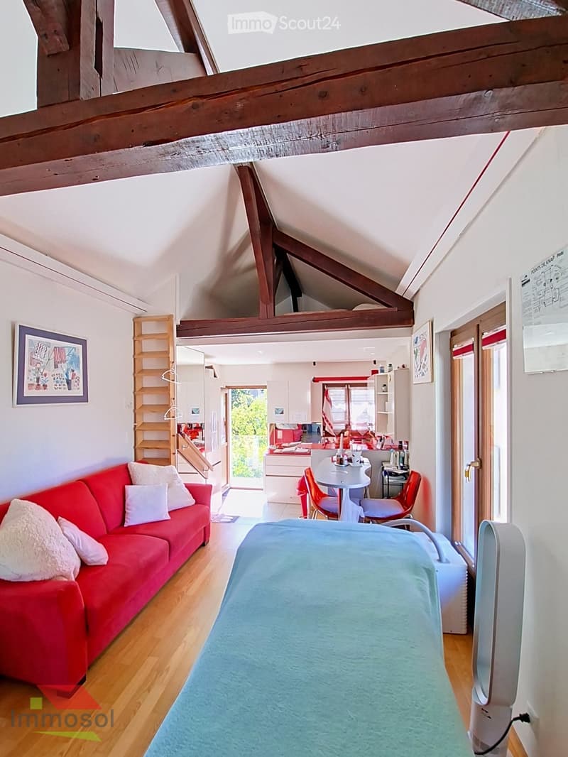 A cozy, loft-style living space with exposed wooden beams, a red sofa, a small dining table with chairs, a staircase leading to an upper level, and a kitchen area visible in the background.
