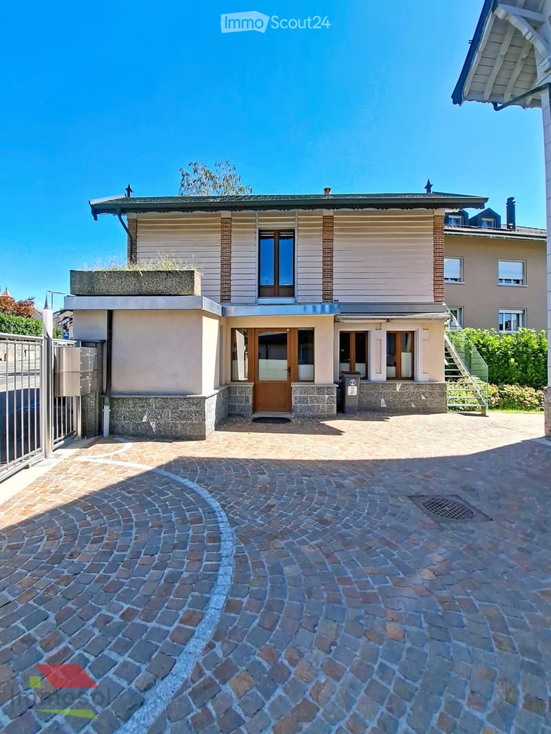 Two-story house with beige siding, brick accents, and a stone foundation. Features a covered entrance with wooden doors and windows, a paved driveway, and a small garden area.