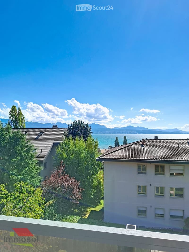 Balcony overlooking a scenic view of a lake and distant mountains, with surrounding greenery and neighboring houses.