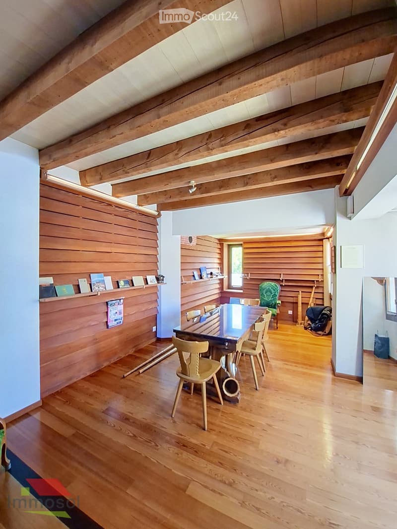 Bright, wooden-floored room with exposed wooden beams and a slanted ceiling. Features a dining table with chairs, a shelf with books and decorative items, and a small window letting in natural light.