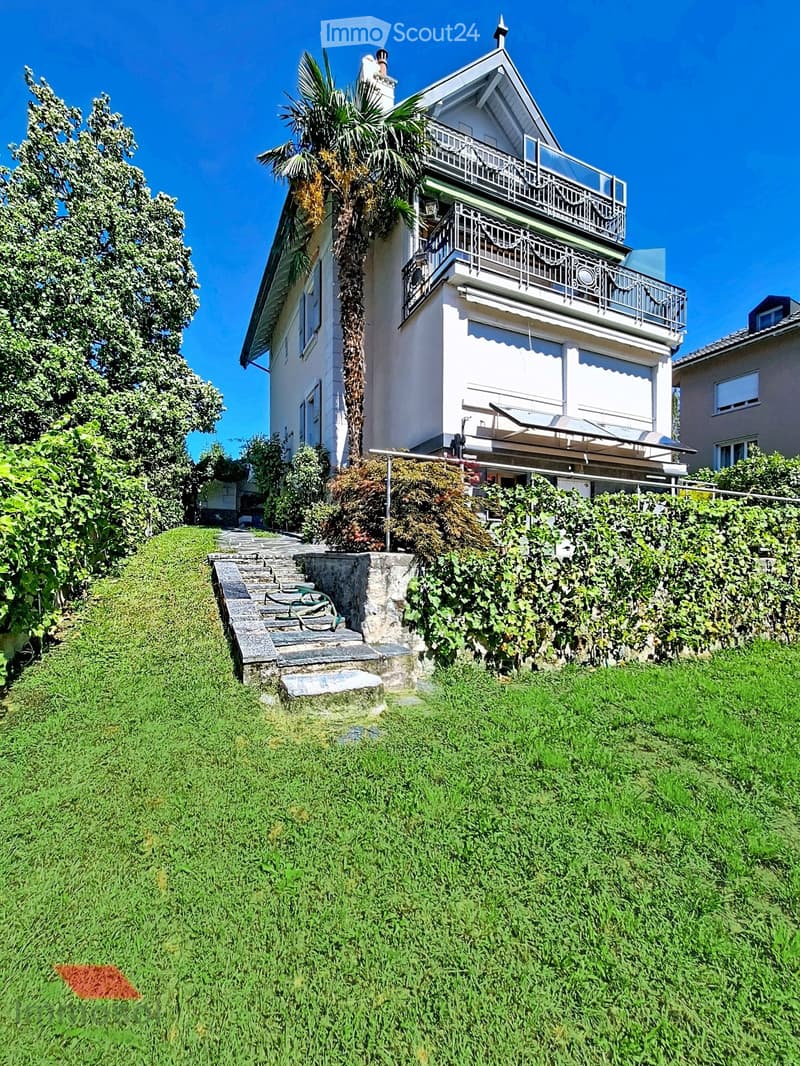 A two-story house with a balcony, surrounded by greenery and a stone staircase leading up to it.