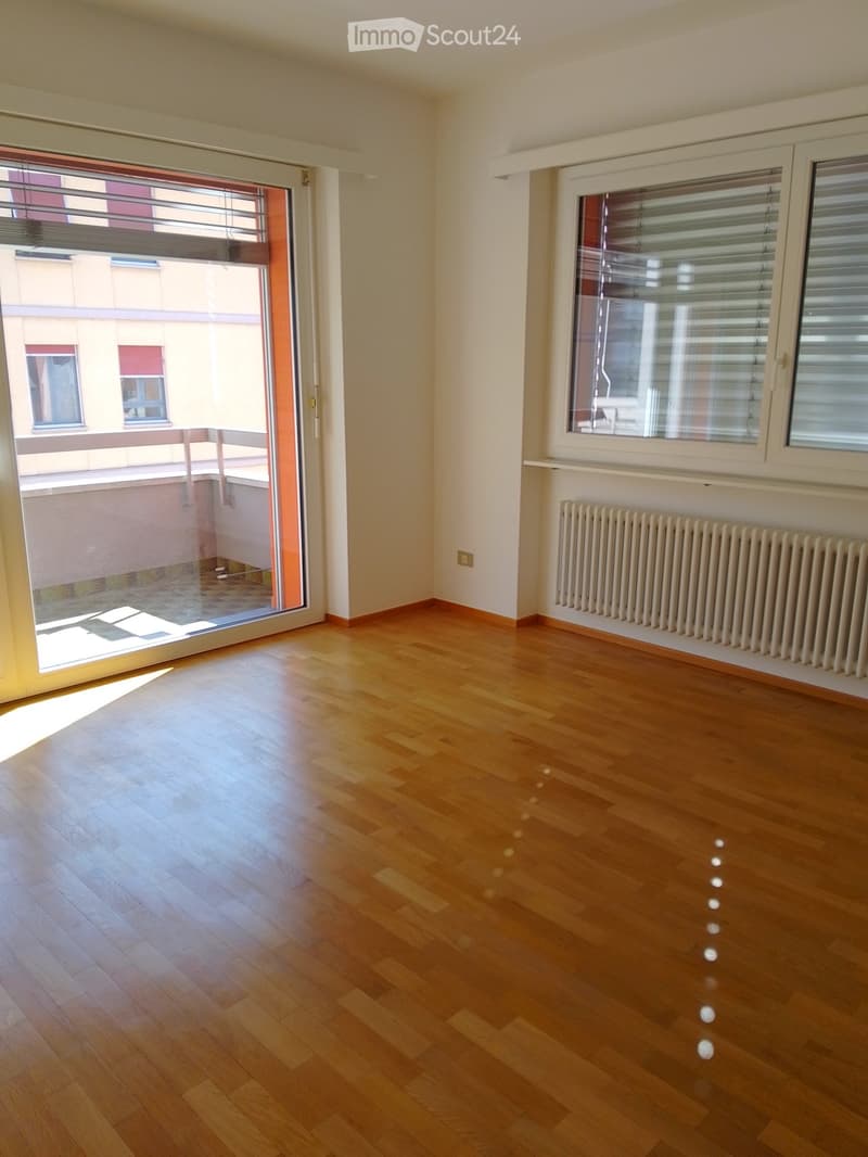 Empty room with wooden flooring, large window with blinds, radiator, and sliding glass door leading to a balcony.