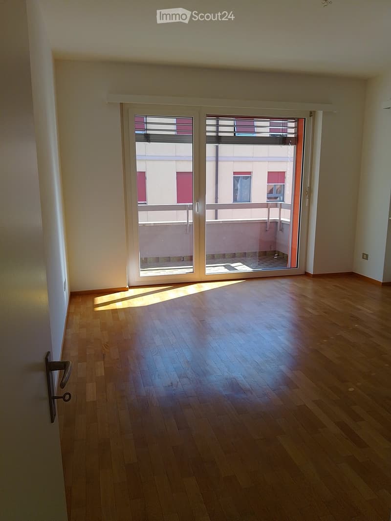Empty room with wooden flooring and large sliding glass doors leading to a balcony. Sunlight streams in, casting shadows on the floor.