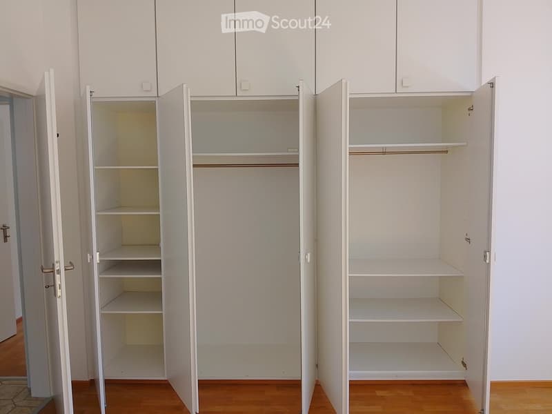 Empty room with white built-in wardrobes and shelves, wooden flooring, and white walls.