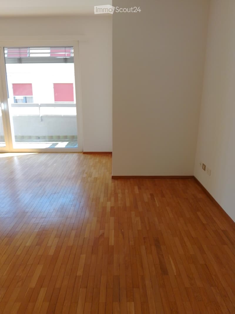 Empty room with wooden flooring and white walls, featuring a large window with a view of a building outside.