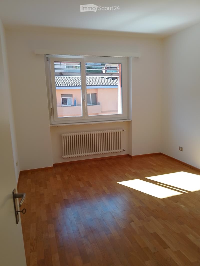Empty room with wooden flooring, white walls, and a radiator below a large window offering a view of neighboring buildings.