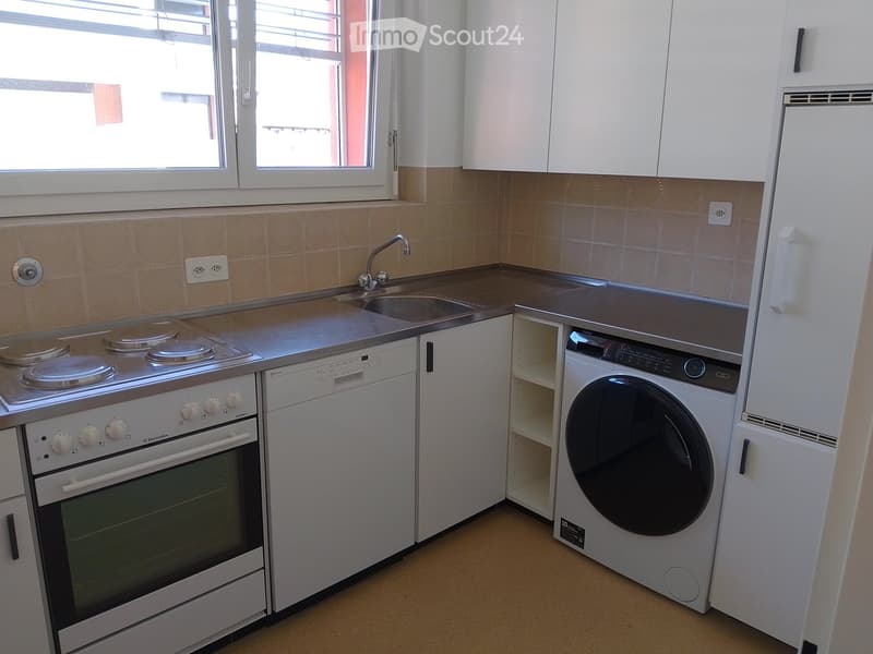 Modern kitchen with white cabinets, stainless steel appliances including an oven, dishwasher, washing machine, and refrigerator. Features a sink with a faucet and a window above.