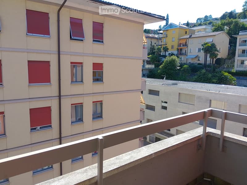 Balcony overlooking a multi-story residential building with closed red shutters and visible windows. The background includes neighboring buildings, greenery, and a clear sky.