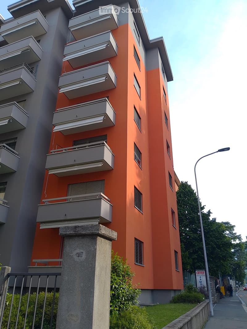 Modern multi-story apartment building with orange and grey facade, multiple balconies, and a raised ground floor entrance.