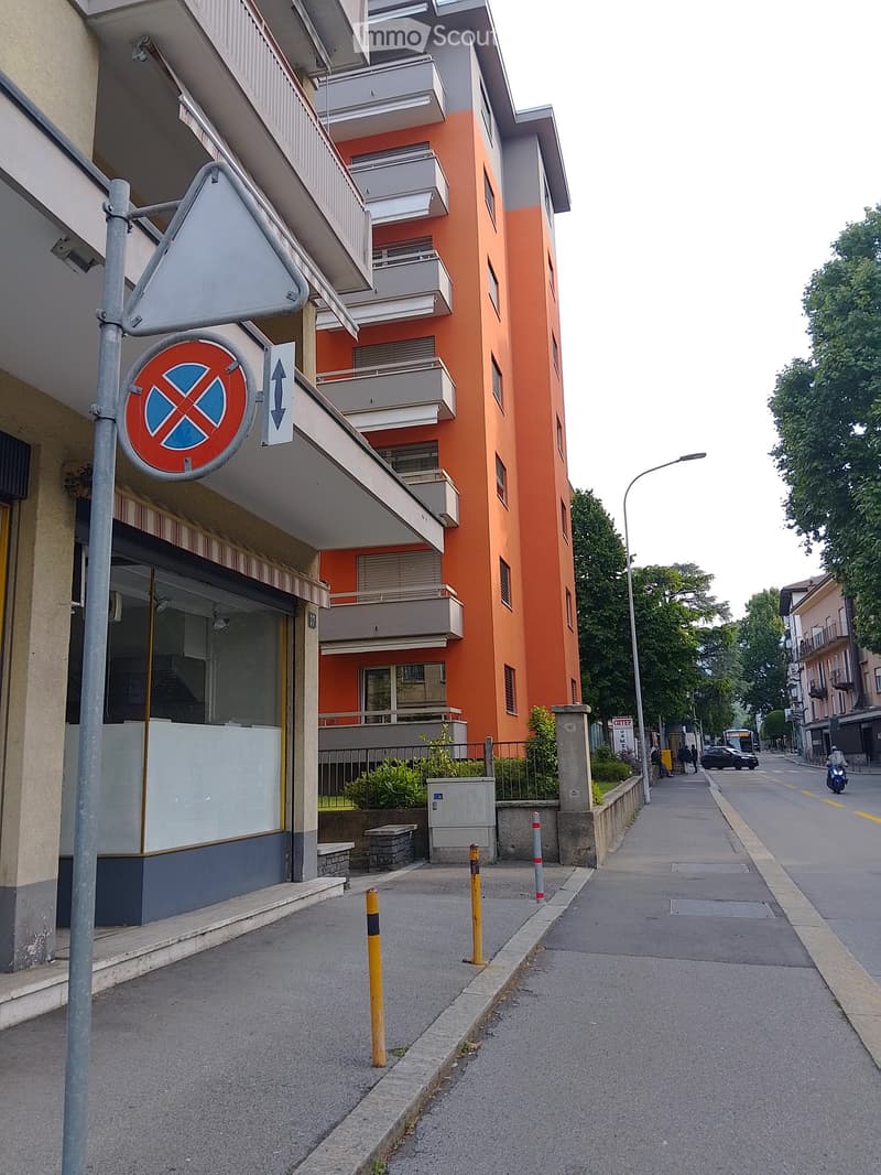 Multi-story residential building with orange and gray facade, balconies, and a storefront on the ground floor. The sidewalk in front has bollards and traffic signs.