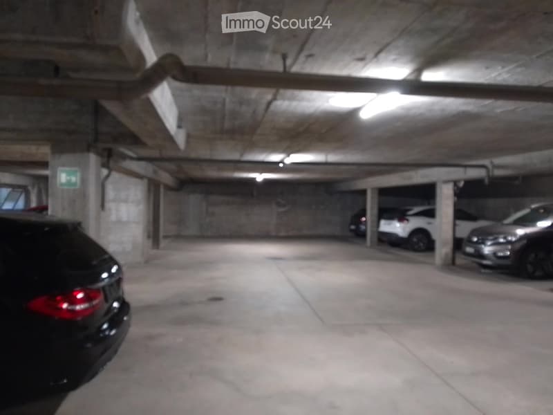 Underground parking area with several parked cars, concrete pillars, and fluorescent lighting.