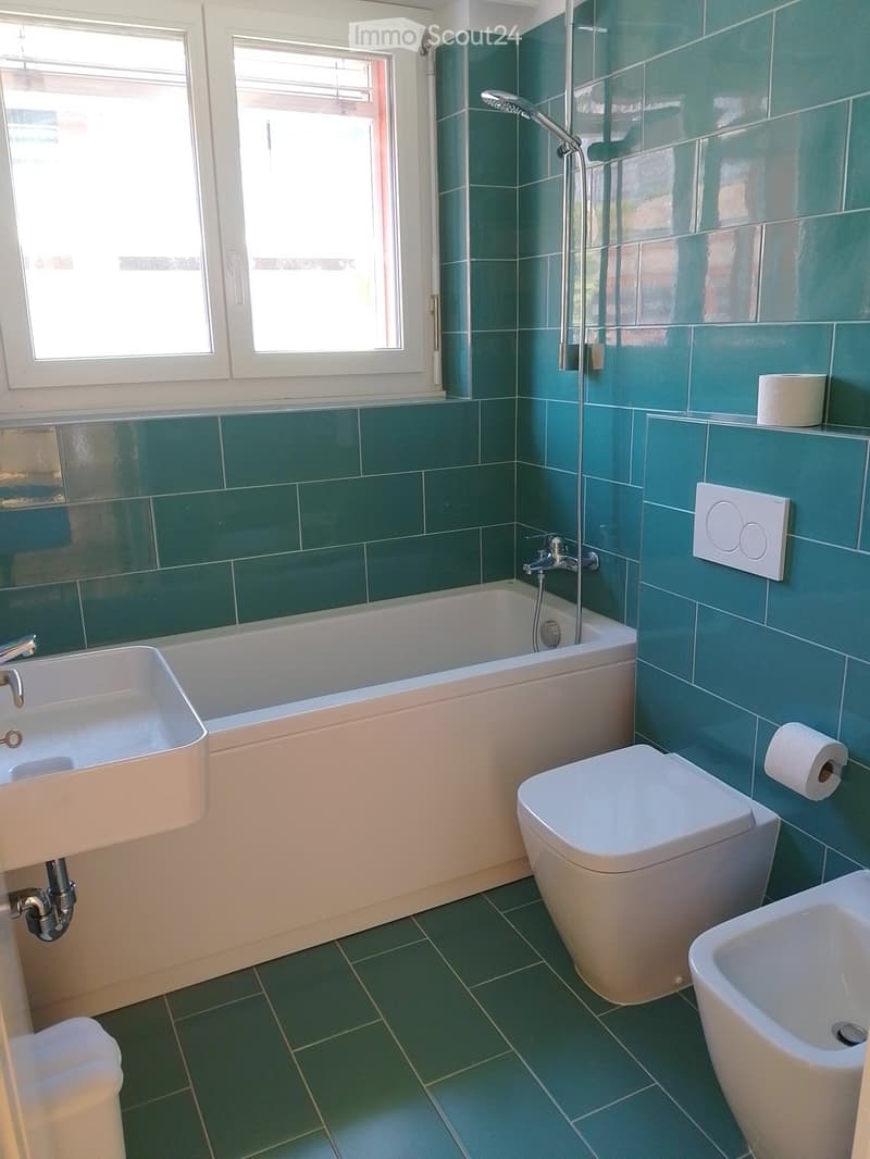 Bathroom with green tiled walls and floor, featuring a bathtub, toilet, sink, and showerhead.