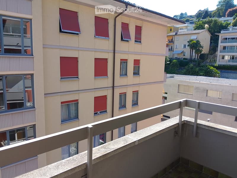 Balcony overlooking a multi-story residential building with red window shutters and a view of neighboring buildings and greenery.