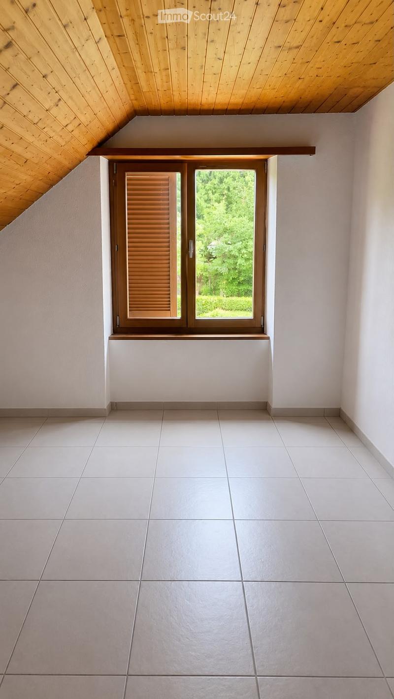 Empty room with white tiled floor, white walls, and a large window with wooden shutters, offering a view of greenery outside.
