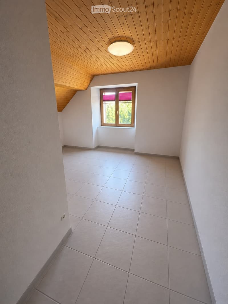 Empty room with tiled floor, white walls, wooden slanted ceiling, and a single window with blinds.