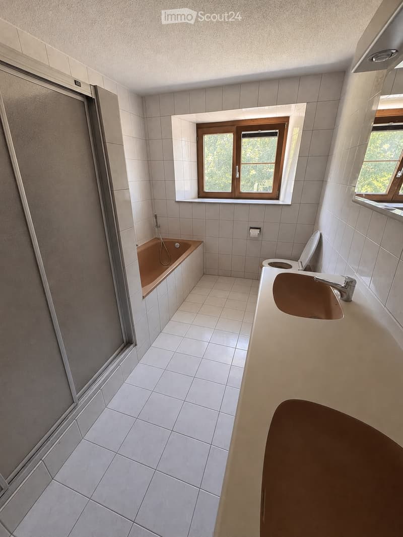 Modern bathroom with white tiled walls and floor, featuring a bathtub, sink with faucet, and toilet. There is a window with wooden frame providing natural light.