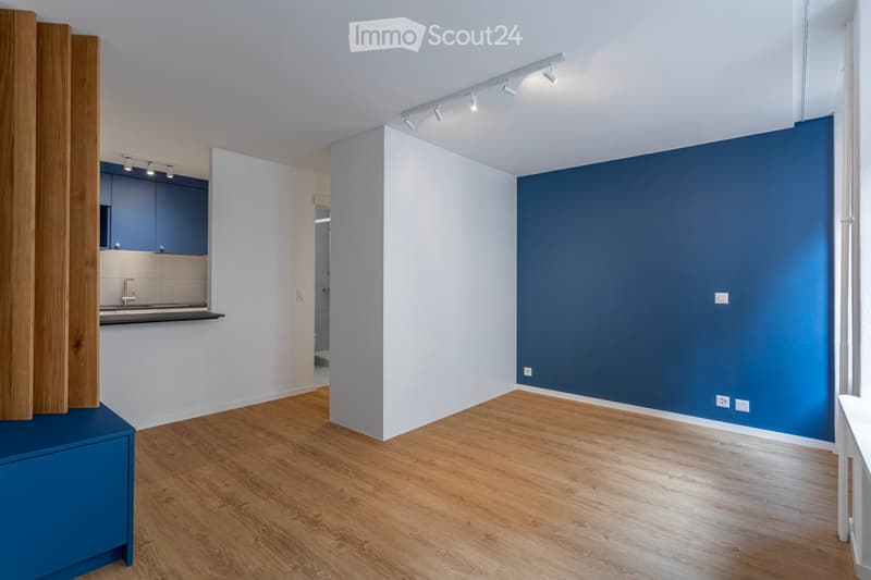 Empty room with wooden flooring, white walls, and a blue accent wall. Features track lighting and a small kitchen area with cabinets and a sink.