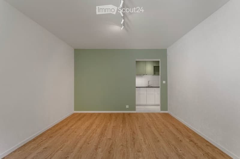 Empty room with wooden flooring, white walls, and a small adjacent kitchen area with white cabinets and a countertop.