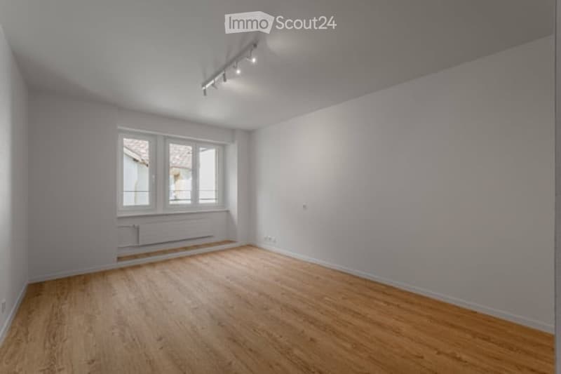 Empty room with wooden flooring, white walls, and large windows allowing natural light. No furniture or equipment is present.