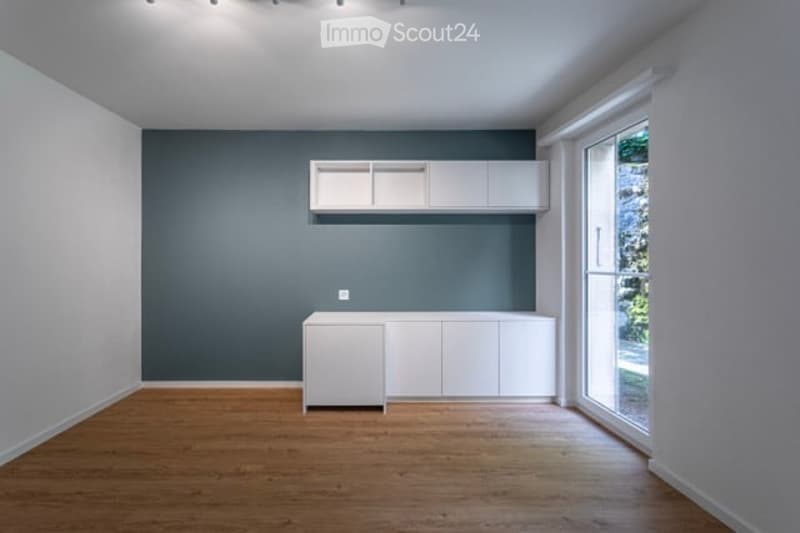 Empty room with white cabinetry, wooden floor, and large window with view to greenery.