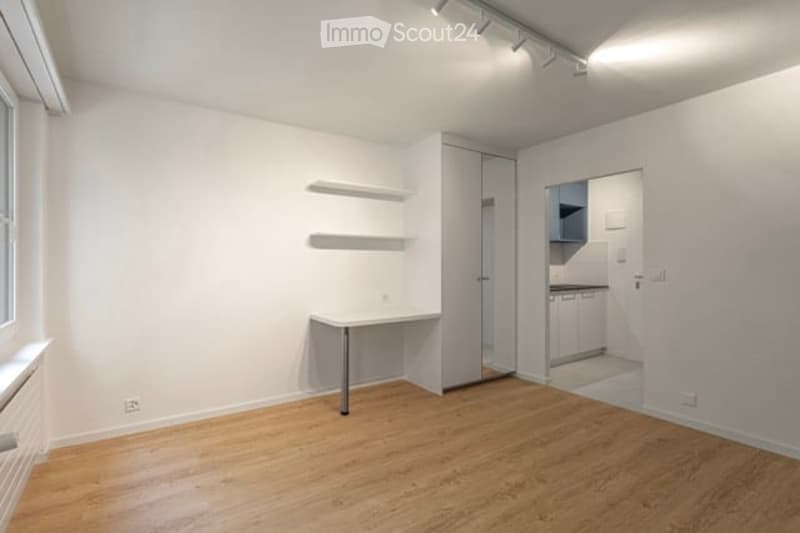 Empty room with wooden flooring, white walls, built-in shelves, and a small table. Adjacent kitchen area visible through an open doorway.