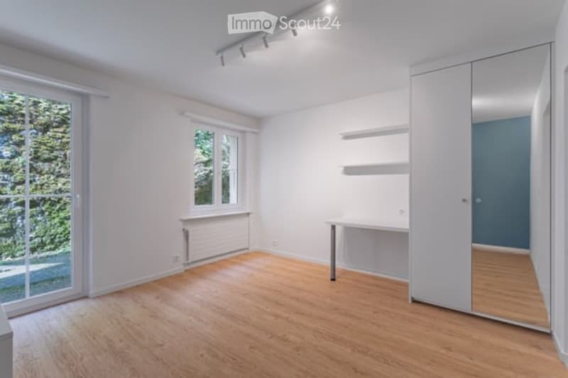 Empty room with light wooden flooring, white walls, large windows, built-in closet with mirrored doors, and a small desk with a chair.