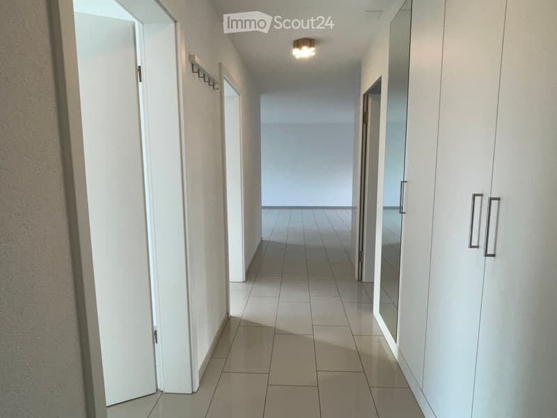A clean, well-lit hallway with tiled flooring, white walls, and several doors. The hallway features a coat rack on one wall and a ceiling light fixture providing illumination.