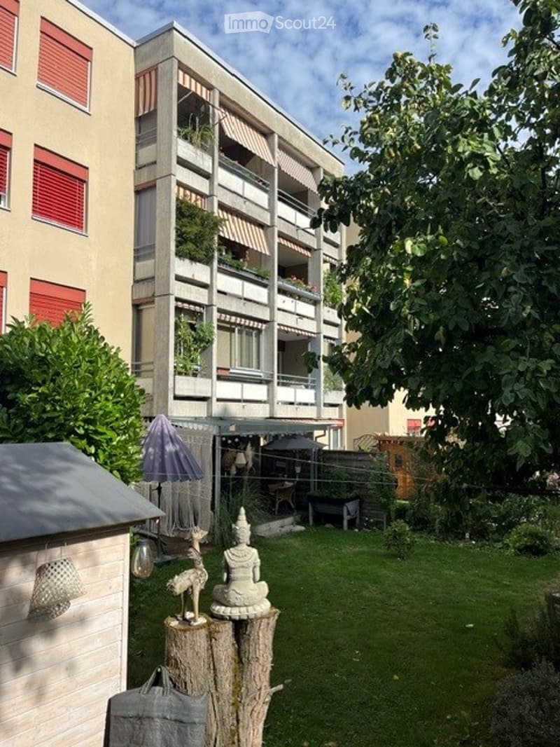 Courtyard garden with a small seating area, a tree stump with a decorative statue, and a small shed. The background shows a multi-story residential building with balconies and greenery.