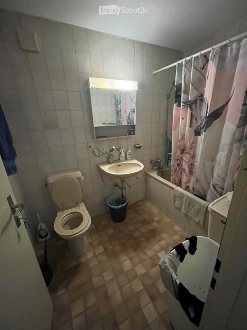 A small bathroom with beige tiles, a toilet, a sink with a mirror above it, a bathtub with a floral shower curtain, and a towel hanging on the side.