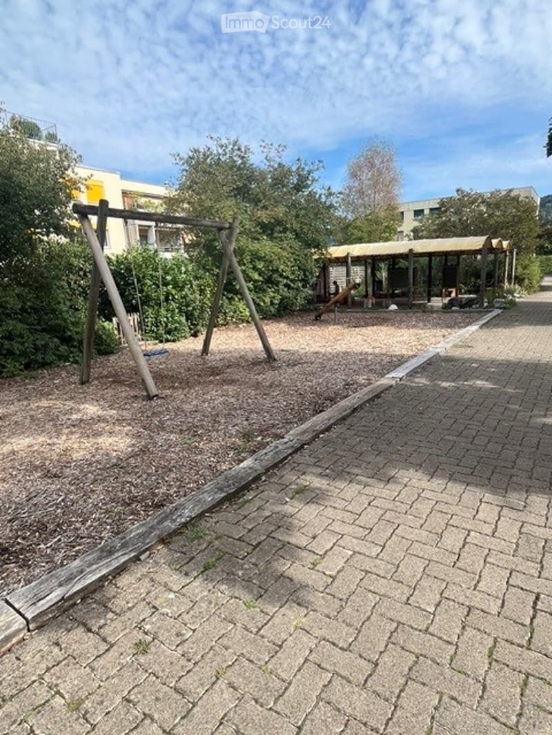 Outdoor playground area with a swing set and a covered seating area, surrounded by trees and a paved walkway.