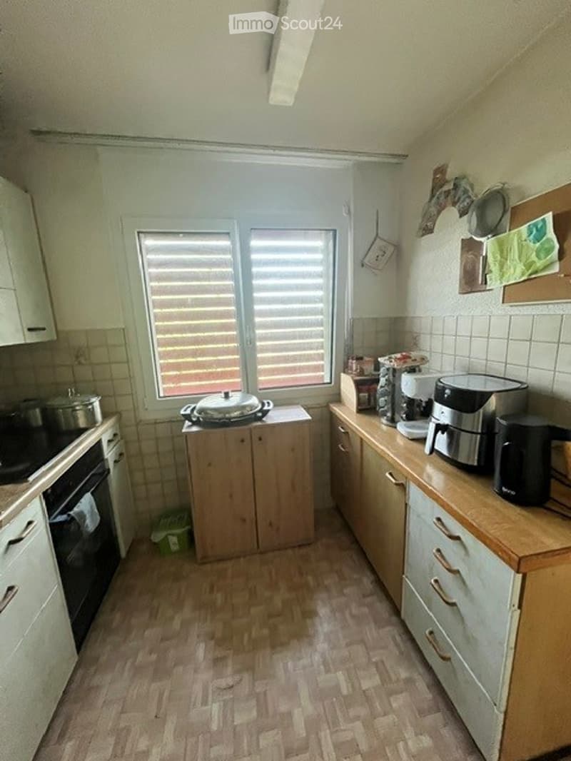 A small kitchen with light wooden cabinets, a window with blinds, an oven, a coffee maker, and a fridge. The floor is made of light wooden tiles.