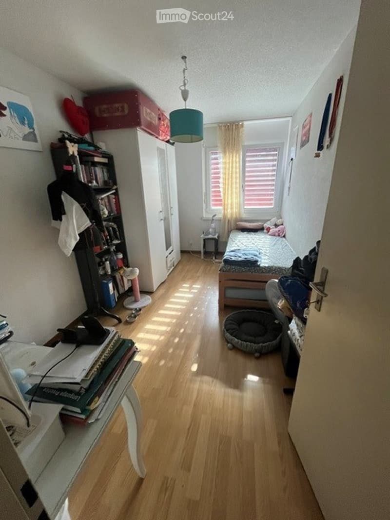 Cozy bedroom with wooden flooring, a bed with pillows and blankets, a wardrobe, a bookshelf, a small stool, a dog bed, a table with books and a chair, and a picture frame on the wall.