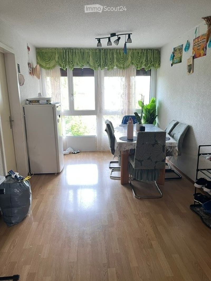 Bright dining area with wooden flooring, large window with green curtains, dining table with chairs, refrigerator, and decorative plant on the table.