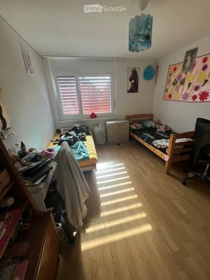 Cozy bedroom with wooden flooring, a single bed, a desk with a chair, and a storage unit. The room features a window with blinds, a colorful wall decoration, and a hanging lamp.