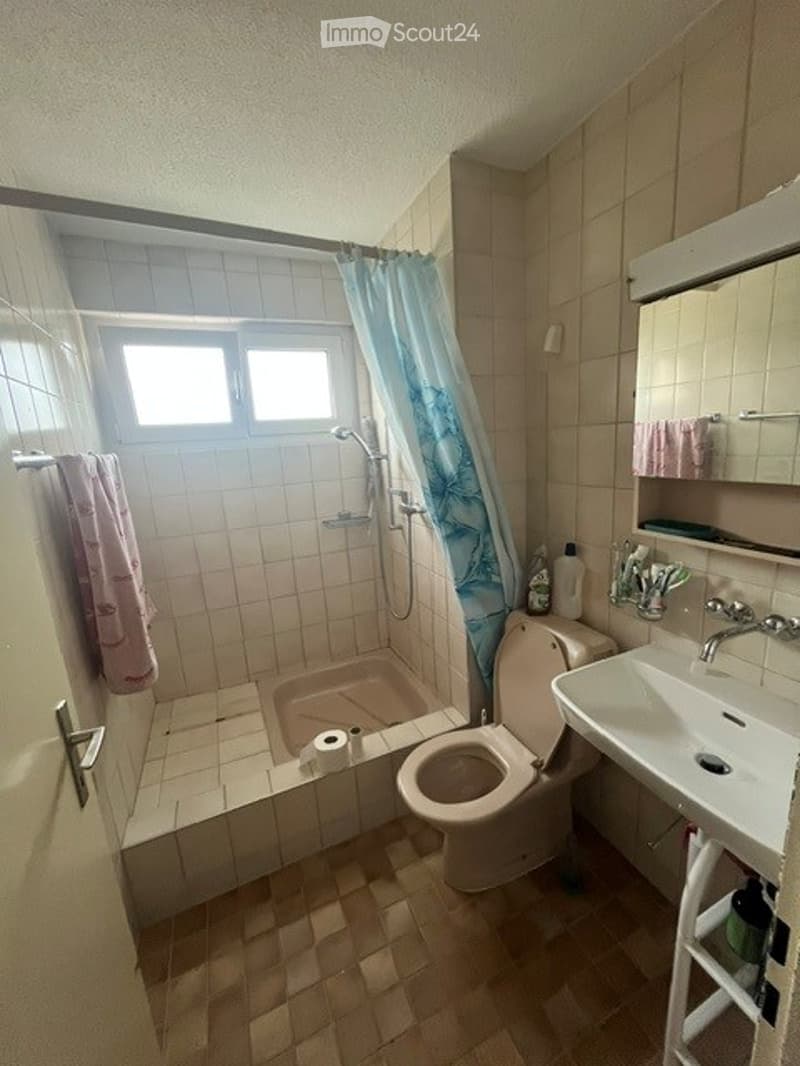 Bathroom with tiled walls and floor, shower area with a blue curtain, toilet, sink, and a small cabinet with toiletries.
