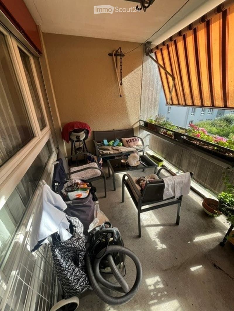Balcony with a small table, chairs, and a baby stroller, with a view of the street and some potted plants.