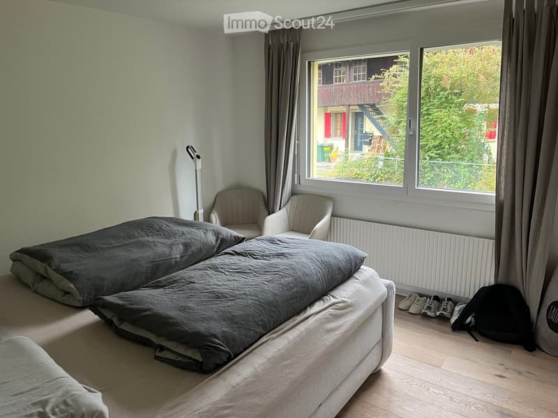 Cozy bedroom with a double bed, two armchairs, large windows with curtains, and a radiator beneath them. Wooden flooring and a view of greenery outside.