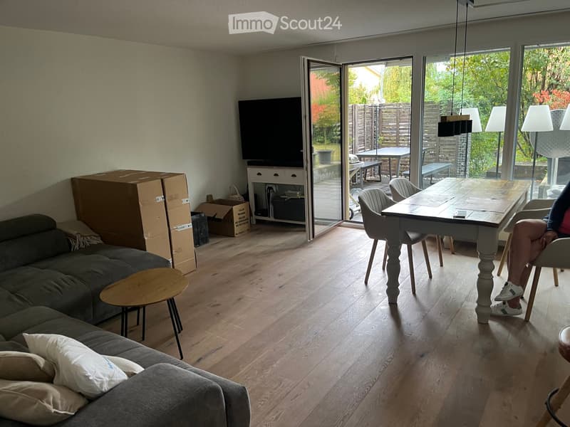 Living room with wooden flooring, grey sofa, coffee table, dining table, and chairs. Large sliding glass doors lead to a patio area with outdoor furniture.