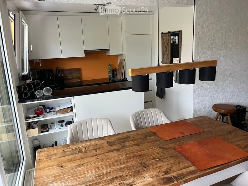 A modern kitchen and dining area with white cabinets, a wooden dining table, two chairs, a stool, and pendant lights. The kitchen counter has various appliances and items, and there are shelves with additional items.