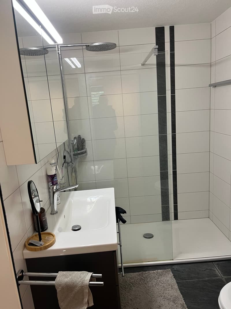 Modern bathroom with a white bathtub, glass-enclosed shower, and a towel on a rack. The shower features a modern design with a glass door and a sleek showerhead. The bathtub has a modern faucet and a small wooden tray with toiletries. A towel is neatly pl