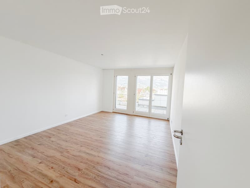 Empty room with light wooden flooring and white walls, featuring large glass doors leading to a balcony with a scenic mountain view.