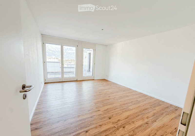 Empty room with light wooden flooring, white walls, and large glass doors leading to a balcony or outdoor area.